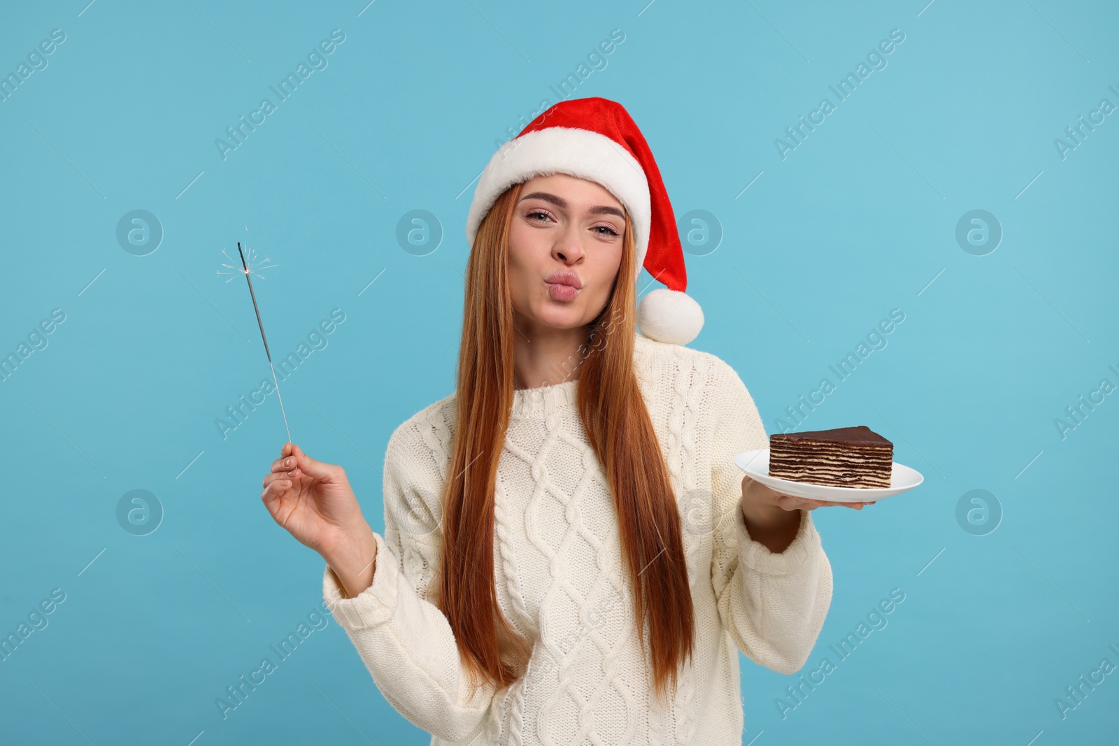 Photo of Young woman in Santa hat with piece of tasty cake and burning sparkler on light blue background