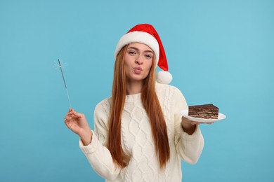 Photo of Young woman in Santa hat with piece of tasty cake and burning sparkler on light blue background