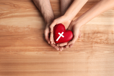 Couple holding heart with cross symbol on wooden background, top view. Christian religion