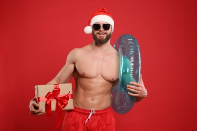 Photo of Muscular young man in Santa hat and sunglasses with inflatable ring and gift box on red background