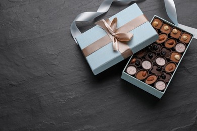 Open box of delicious chocolate candies and light blue ribbon on black table, flat lay. Space for text