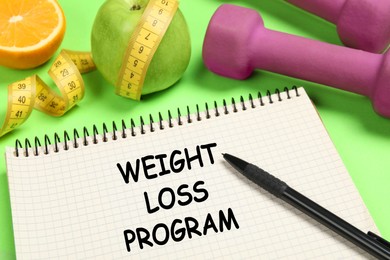 Photo of Weight loss concept. Notebook, dumbbells and apple with measuring tape on green background, closeup
