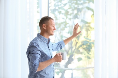Photo of Young man standing near window with open curtains at home