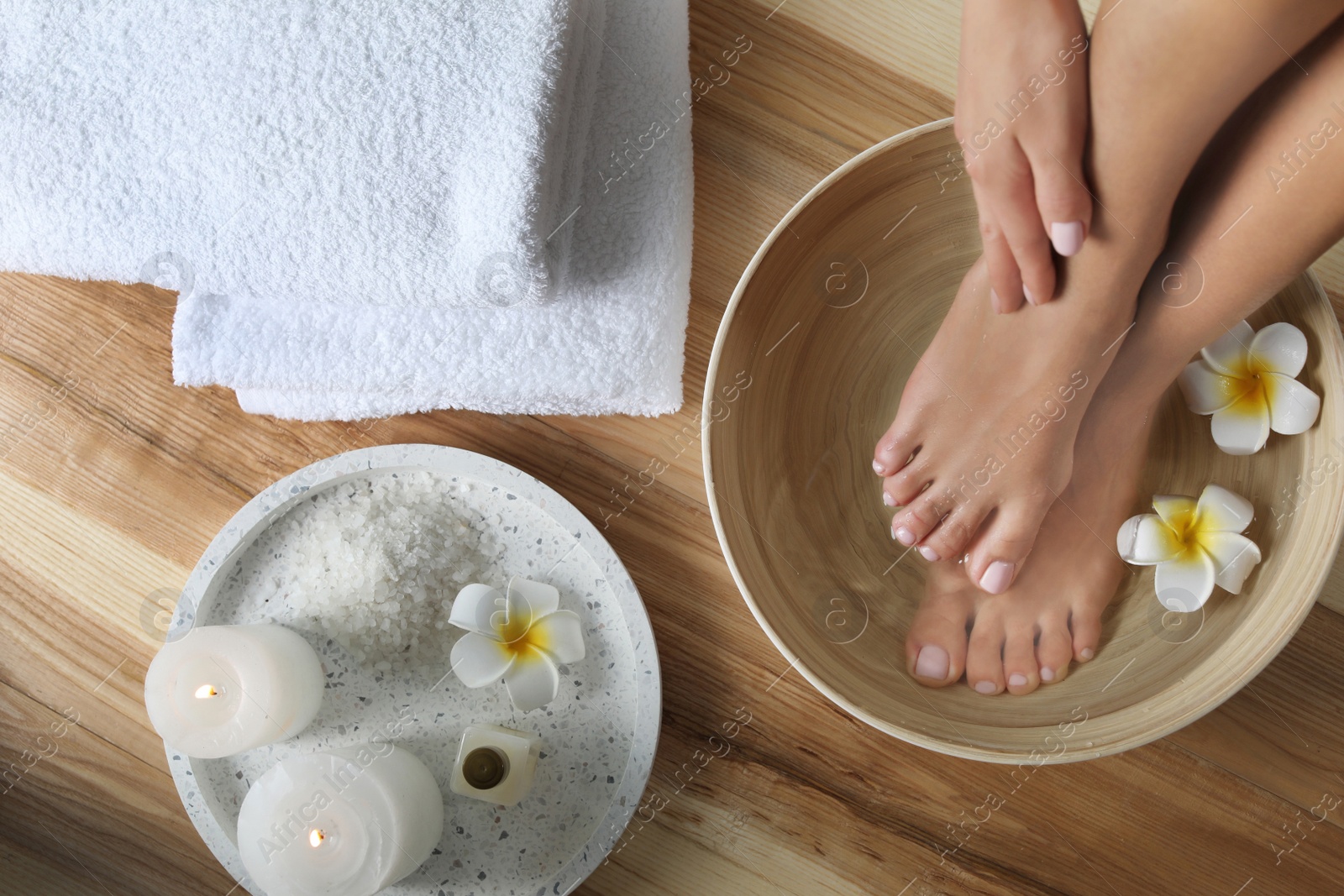 Photo of Woman soaking her feet in dish with water and flowers on wooden floor, top view. Spa treatment
