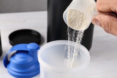 Man pouring protein powder from measuring scoop into shaker on white table, closeup