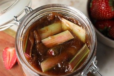 Jar of tasty rhubarb jam and fresh strawberries on table, closeup