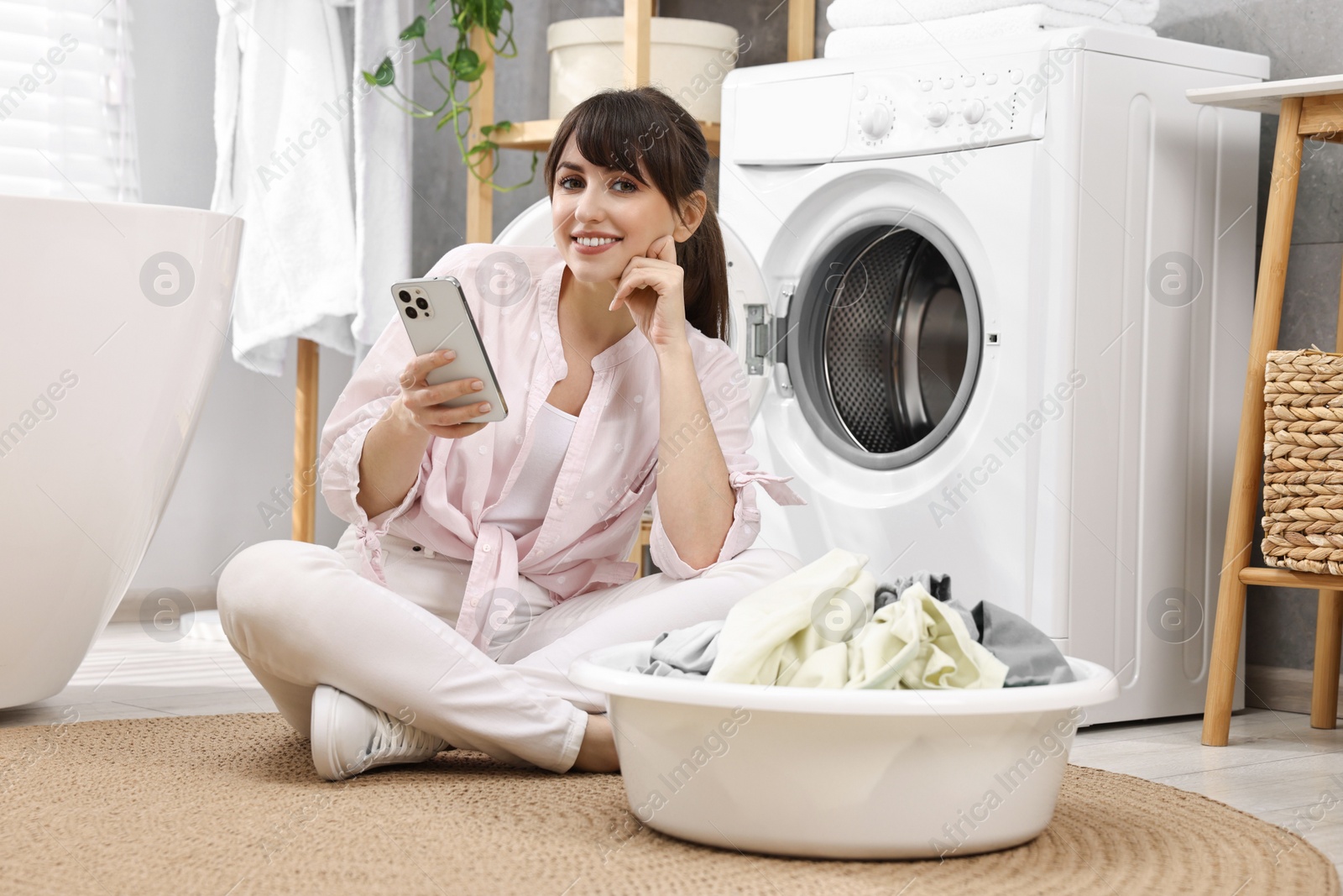 Photo of Happy young housewife with laundry using smartphone near washing machine at home