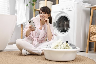 Happy young housewife with laundry using smartphone near washing machine at home