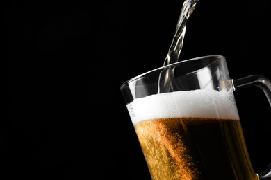 Photo of Pouring cold tasty beer into glass mug on black background, closeup. Space for text