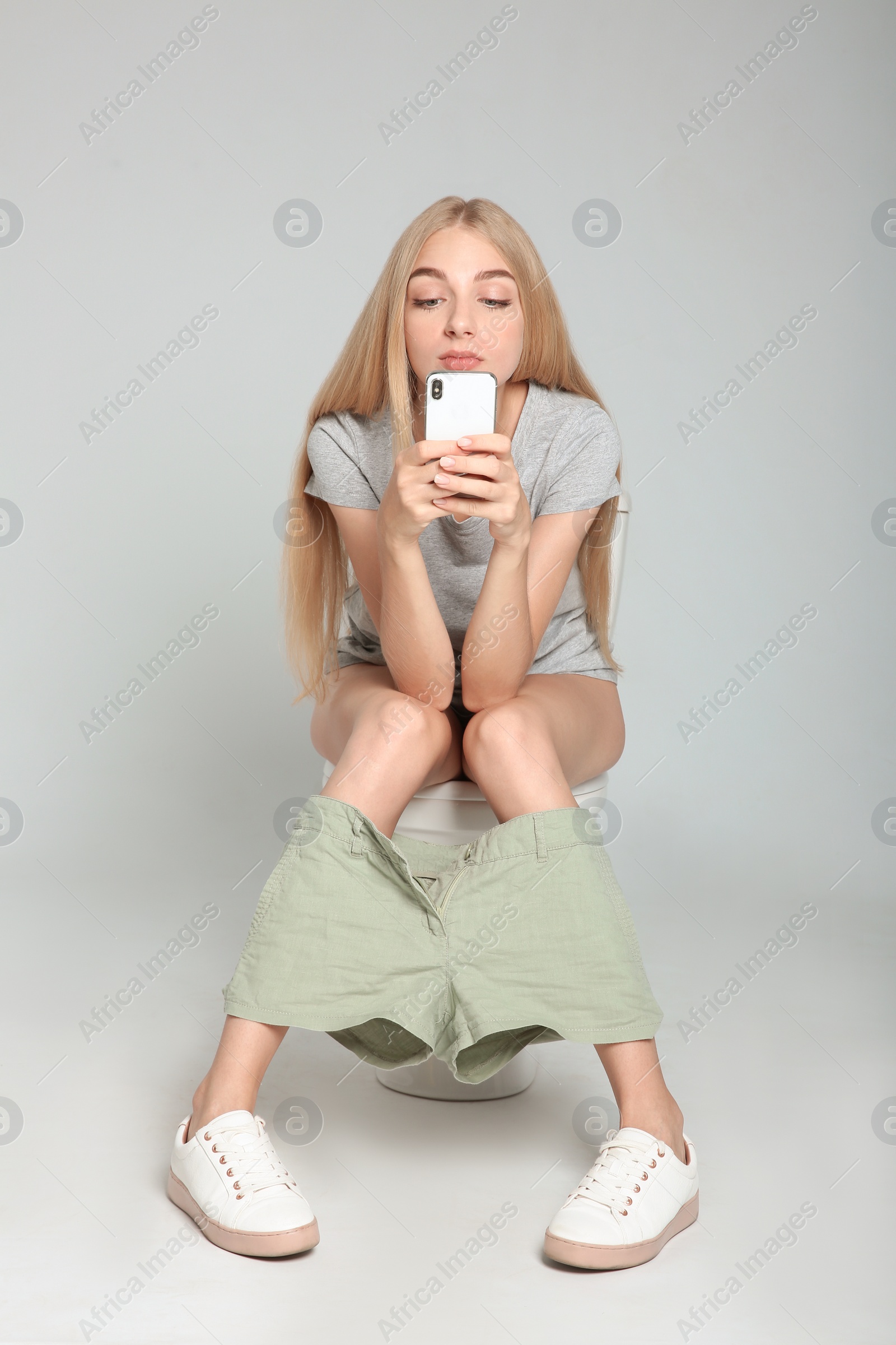 Photo of Young woman using mobile phone while sitting on toilet bowl against gray background
