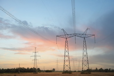 Photo of High voltage towers with electricity transmission power lines in field at sunset