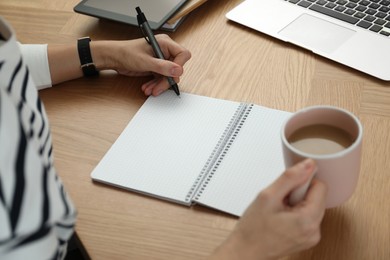 Photo of Left-handed woman with cup of coffee writing in notebook at wooden table, closeup