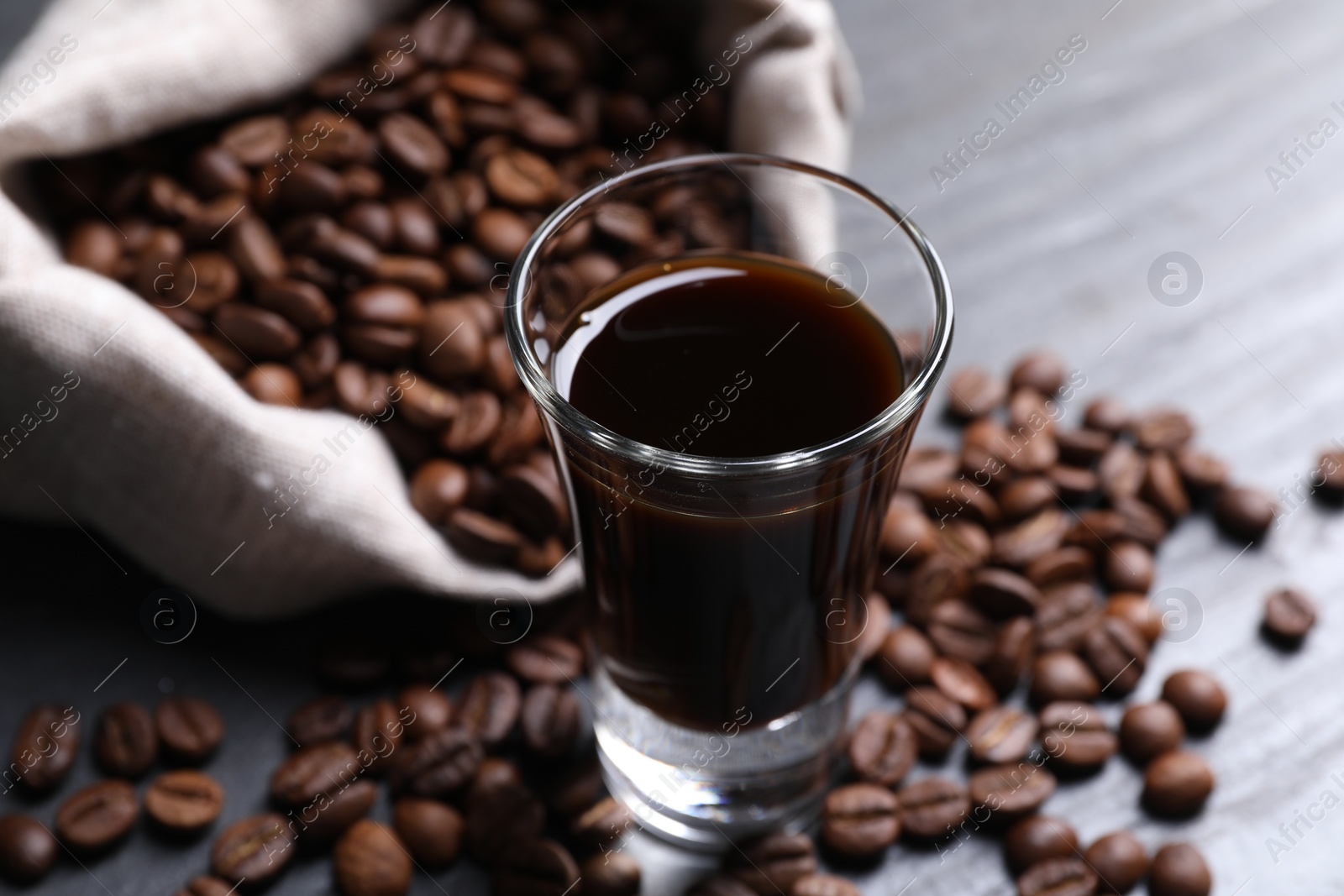 Photo of Shot glass of coffee liqueur and beans on grey table, closeup