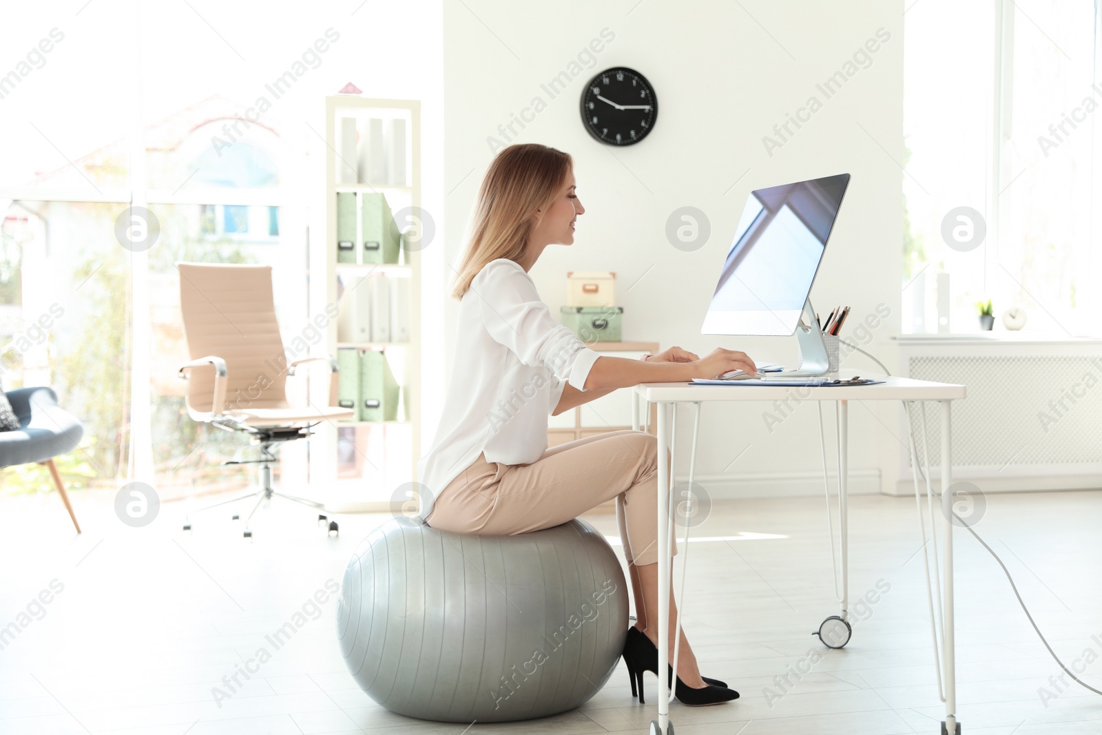 Photo of Young businesswoman sitting on fitness ball and using computer in office. Workplace exercises