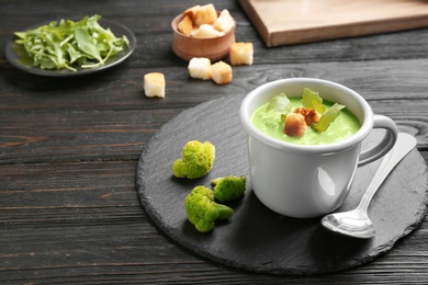 Photo of Cup of broccoli cream soup with croutons served on black wooden table, space for text