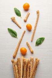 Delicious grissini sticks, basil leaves and yellow tomatoes on white wooden table, flat lay