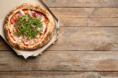 Photo of Tasty pizza with meat and arugula in cardboard box on wooden table, top view. Space for text