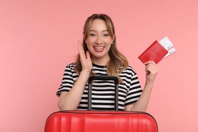 Happy young woman with passport, ticket and suitcase on pink background