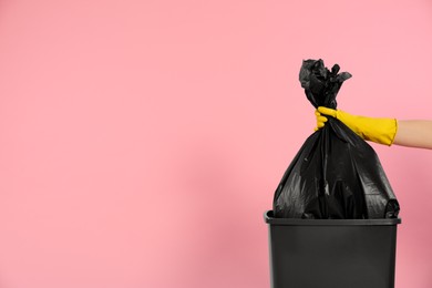 Photo of Janitor in rubber glove holding trash bag full of garbage over bucket on pink background, closeup. Space for text