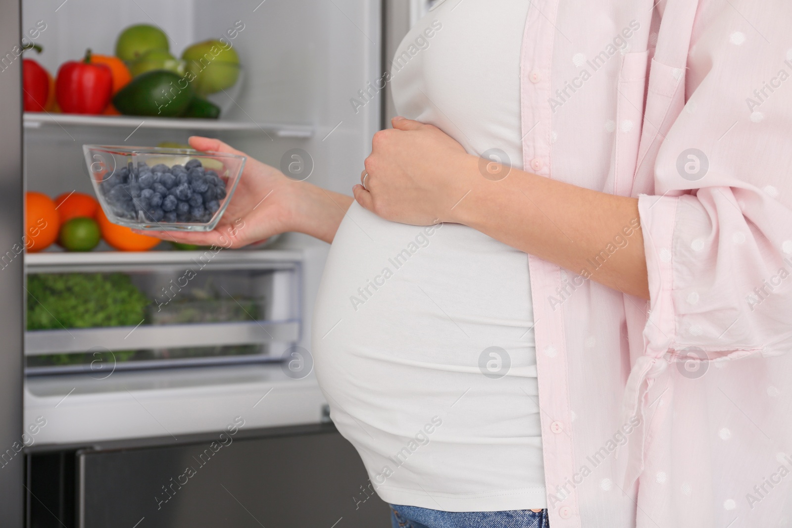 Photo of Young pregnant woman with blueberries near fridge at home, closeup. Healthy eating