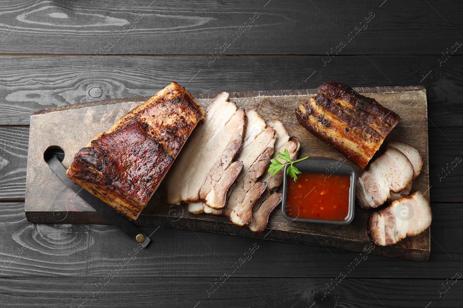 Photo of Pieces of baked pork belly served with sauce and parsley on black wooden table, top view