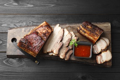 Photo of Pieces of baked pork belly served with sauce and parsley on black wooden table, top view