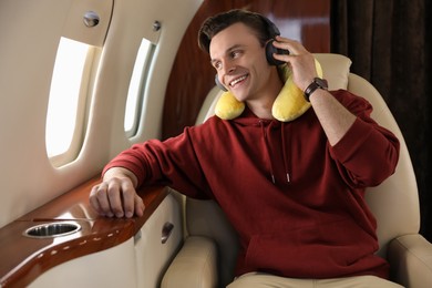 Photo of Young man with travel pillow and headphones listening to music in airplane during flight