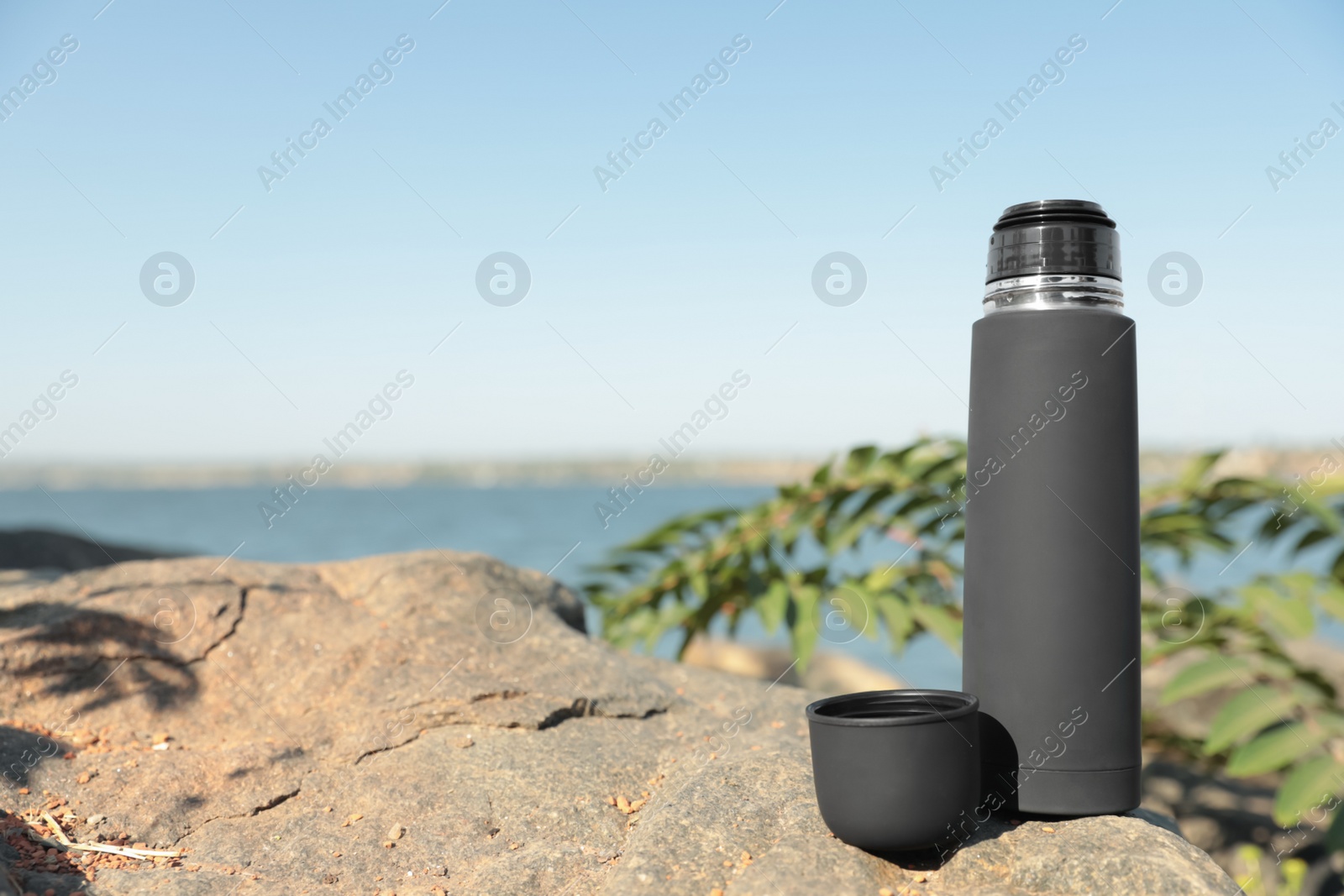 Photo of Modern black thermos on stone near river. Space for text