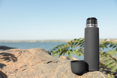 Modern black thermos on stone near river. Space for text