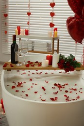 Photo of Wooden tray with wine, burning candles and rose petals on tub in bathroom. Valentine's day celebration