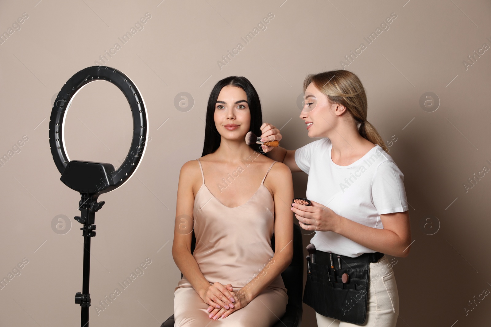 Photo of Professional makeup artist working with beautiful young woman against beige background. Using ring lamp