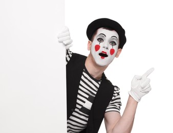 Photo of Funny mime artist peeking out of blank poster and pointing at something on white background