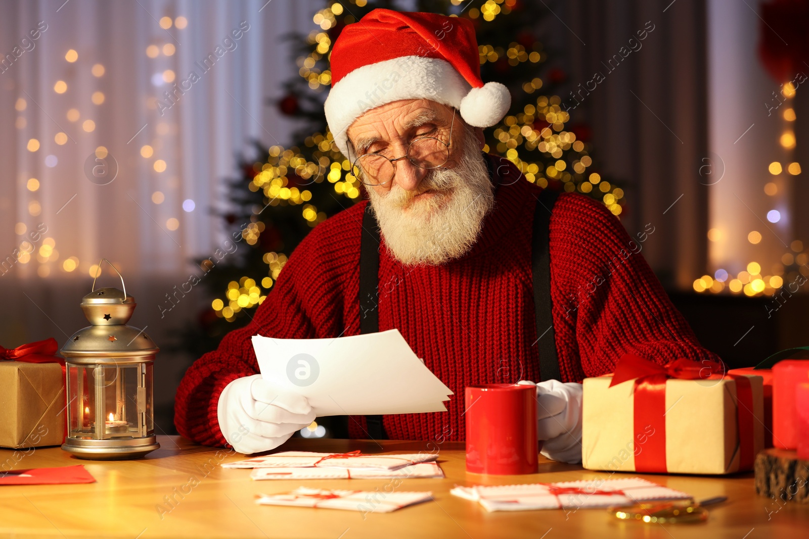 Photo of Santa Claus reading letter and drinking hot beverage at his workplace in room decorated for Christmas