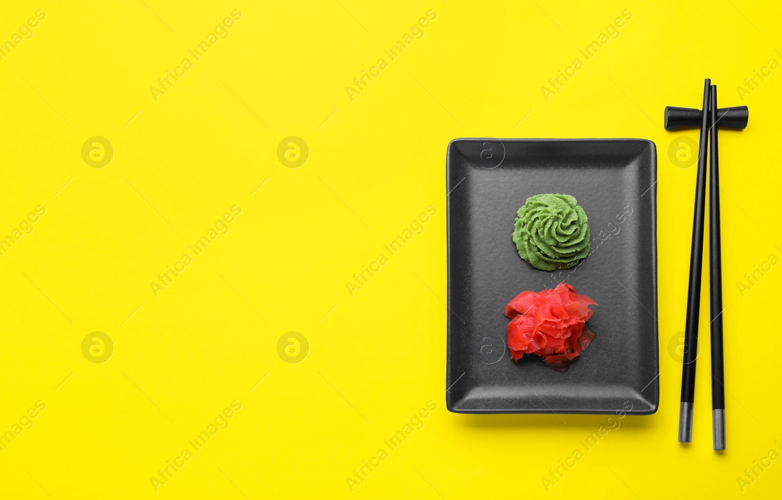 Photo of Chopsticks near plate with swirl of wasabi paste and pickled ginger on yellow background, flat lay. Space for text