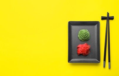 Chopsticks near plate with swirl of wasabi paste and pickled ginger on yellow background, flat lay. Space for text