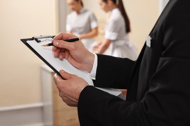 Photo of Man wearing suit with clipboard checking maid's work in hotel room, closeup. Professional butler courses