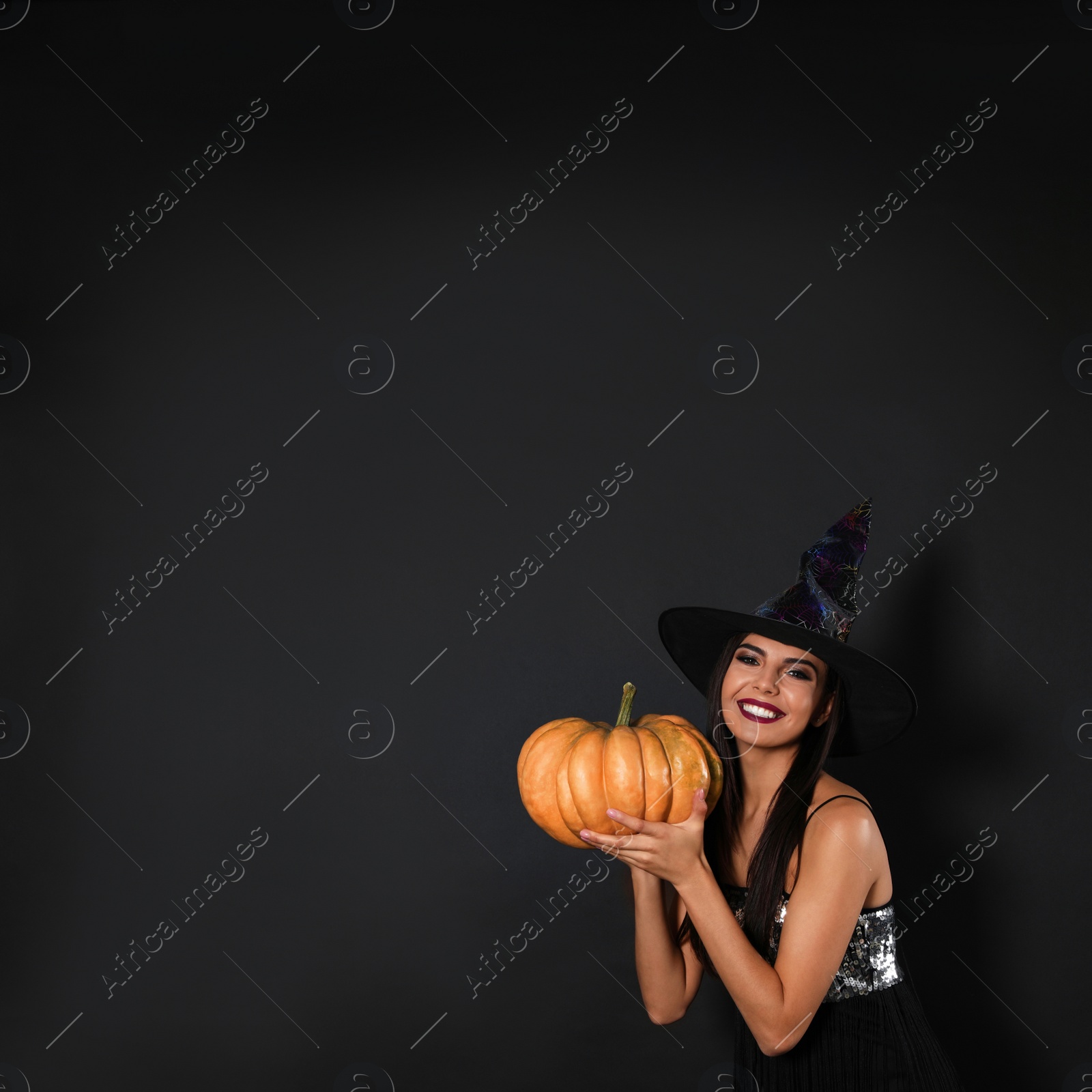 Photo of Beautiful woman wearing witch costume with pumpkin for Halloween party on black background, space for text