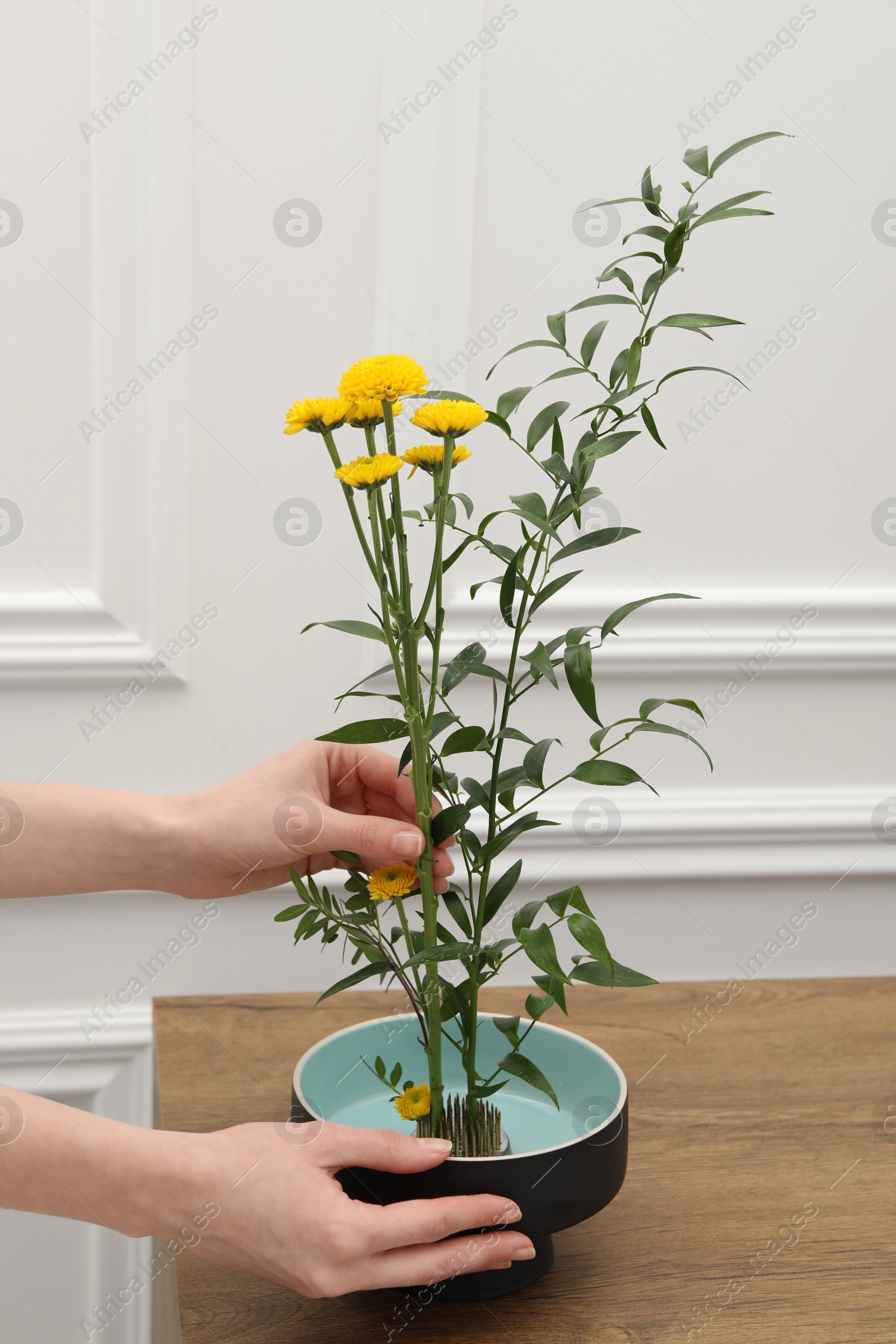Photo of Woman creating stylish ikebana with beautiful yellow flowers and green branch at wooden table, closeup