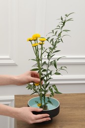 Photo of Woman creating stylish ikebana with beautiful yellow flowers and green branch at wooden table, closeup