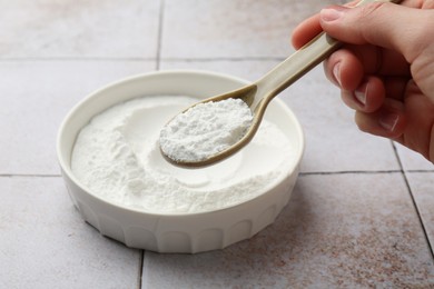 Woman taking baking powder with spoon from bowl at light tiled table, closeup