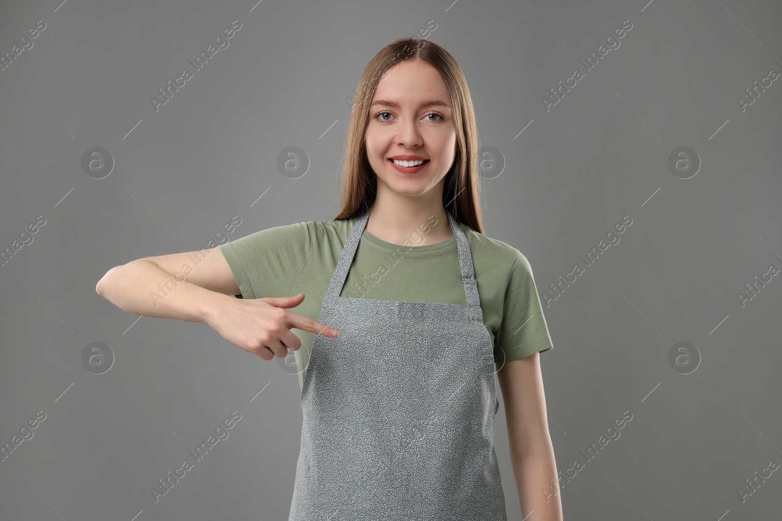 Photo of Beautiful young woman pointing at kitchen apron on grey background. Mockup for design