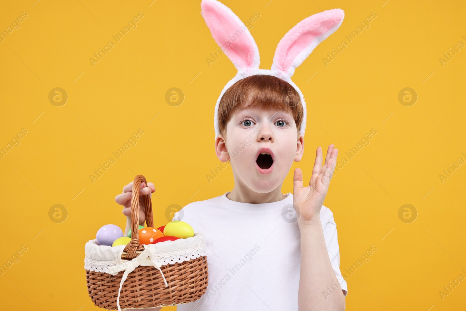 Photo of Easter celebration. Surprised little boy with bunny ears and wicker basket full of painted eggs on orange background