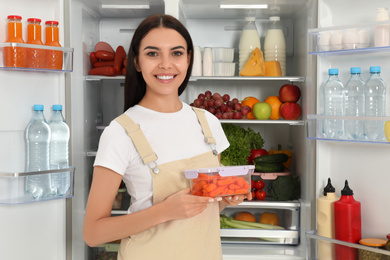 Photo of Young woman with lunchbox of carrots near refrigerator