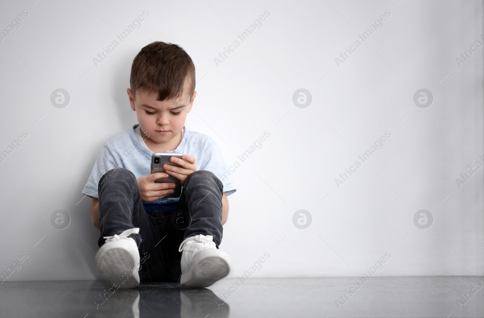 Photo of Sad little boy with mobile phone sitting  near white wall, space for text