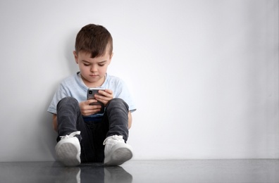 Photo of Sad little boy with mobile phone sitting  near white wall, space for text