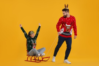 Photo of Young couple in Christmas sweaters. Man pulling his woman in sled on orange background