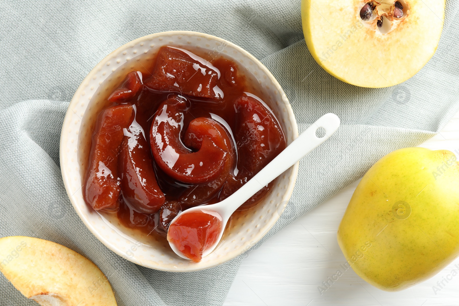 Photo of Tasty homemade quince jam in bowl, spoon and fruits on white wooden table, flat lay