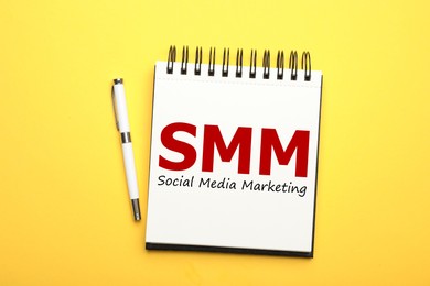 Notebook with text SMM (Social media marketing) and pen on yellow background, flat lay 