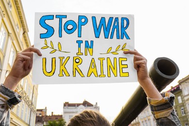 Photo of Woman holding poster in colors of national flag with words Stop War In Ukraine near broken military tank on city street, closeup