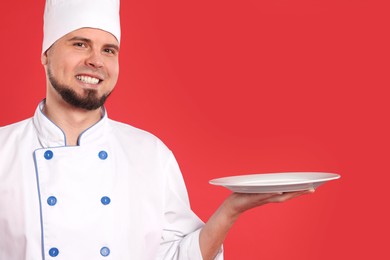 Photo of Happy professional confectioner in uniform holding empty plate on red background. Space for text
